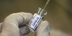 ein Hand in einem Gummihandschuh hält ein kleines Fläschchen in dem eine Pipette steckt, auf dem Fläschchen steht "Covid-19 mRNA Vaccine"