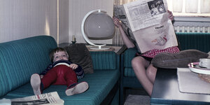 Auf einen historischen Bild liegt ein Kind auf dem Sofa, während eine Frau in einer Zeitung liest.