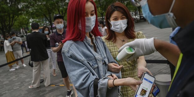 Zwie Frauen mit Mundschutz, eine trägt ein Smartphone in der Hand, stehenm vor einem Mann der die Körpertemperatur misst