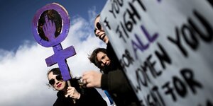 eine Frau trägt auf der Frauendemo ein lila Frauenzeichen mit Stinkefinger