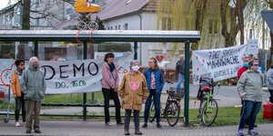 Menschen stehen in Abstand an einer Bushaltestelle, auf der Haltestelle steht ein Kinderwagen, and er Haltestelle hängt ein Plakat auf dem steht: Demo 16.04.
