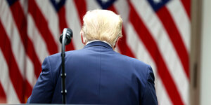 Der Rücken und Hinterkopf von Donald Trump vor der US-Fahne, vor seinem Rücken ein Mikrofonständer