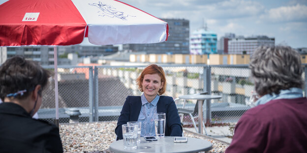 Katja Kipping, eine Frau mit kinnlangen hellen Haaren sitzt an einem Tisch auf der Dachterasse der taz. Im Hintergrund sind Häuser zu sehen. Über Kipping ein roter Sonnenschirm. Vor Kipping zwei Menschen.
