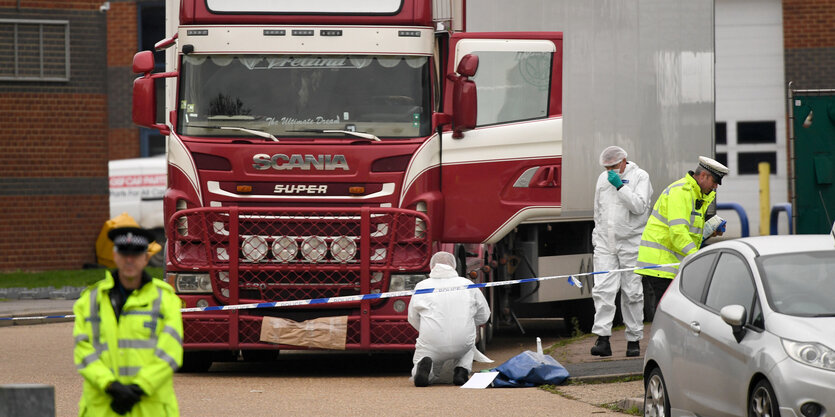 Der Lastwagen, in dem 39 Geflüchtete starben. Das Führerhaus ist rot, der Rest weiß. Ein Polizist in Warnjacke hält Wache. Zwei Kriminalbeamte in weißen Ganzkörperanzügen sammeln spuren.