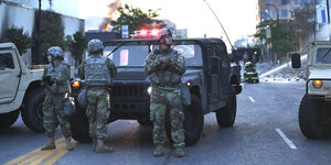 Bewaffnete Uniformierte sind vor einem Militärfahrzeug postiert. Es ist früher Morgen, im Hintergrund löschen Feuerwehrleute einen Brand