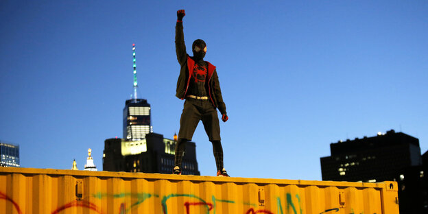 Ein Mann mit Spiderman-Kostüm steht mit erhobener Faust auf einem Container.