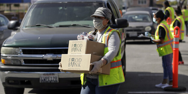 Eine Frau in gelber Warnweste und mit Mundschutz trägt Esssens-Kartons über eine Straße mit vielen Autos