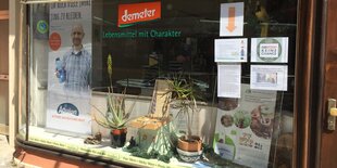 Ein Schaufenster mit Demeterlogo, daneben hängen beschriebene Din A-4-Seiten