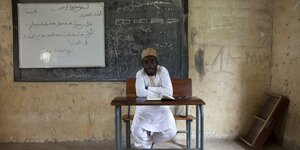 Ein Mann sitzt an einem kleinen Tisch vor einer Tafel. er ist ein Lehrer an einer islamischen Schule in Kaduna in Nigeria. Er trägt weißes Gewand und einen Hut.