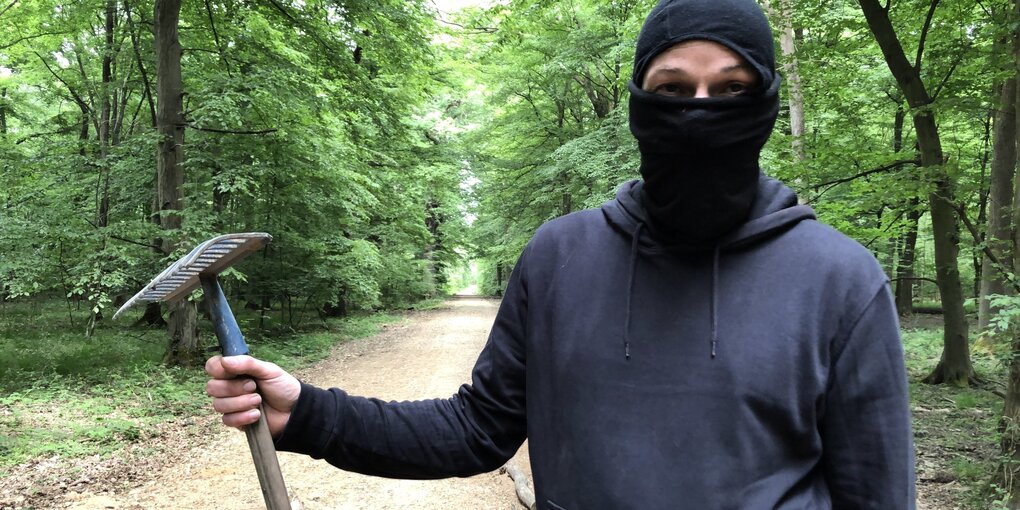 Eine vermummte Person steht mit einem Rechen im Wald.