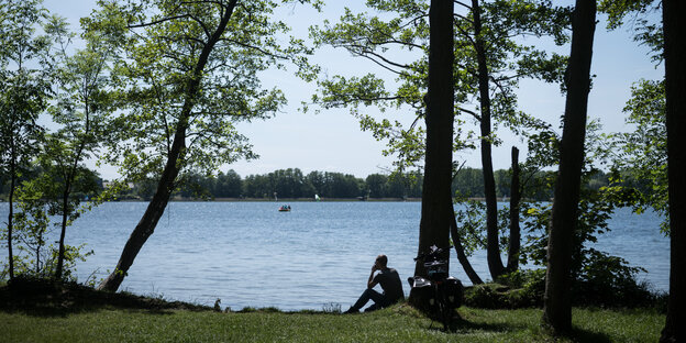 Ein Mann sitzt an einem See unter einem Baum. Neben ihm steht ein Fahrrad. Auf dem See sind ein kleines Boot und mehrere Segel.