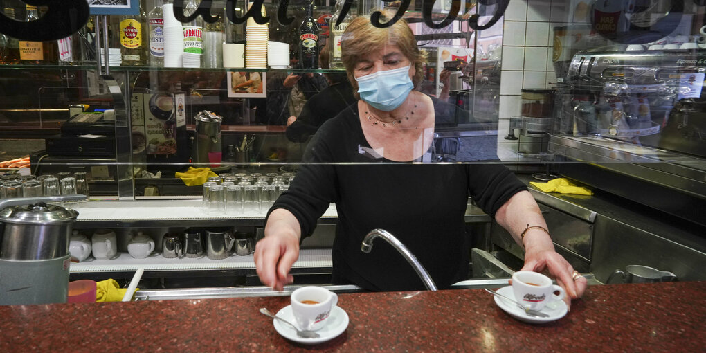 Eine Frau mit Mundschutz reicht zwei Kaffeetassen unter einer Plexiglasscheibe durch.