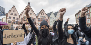Junge Frauen demonstrieren. Sie heben ihre Fäuste in die Luft und haben einen Karton dabei, auf dem Black Lives Matter steht. Die Frauen tragen Mundschutz oder Schal vor dem Mund. Im Hintergrund stehen alte Häuser.