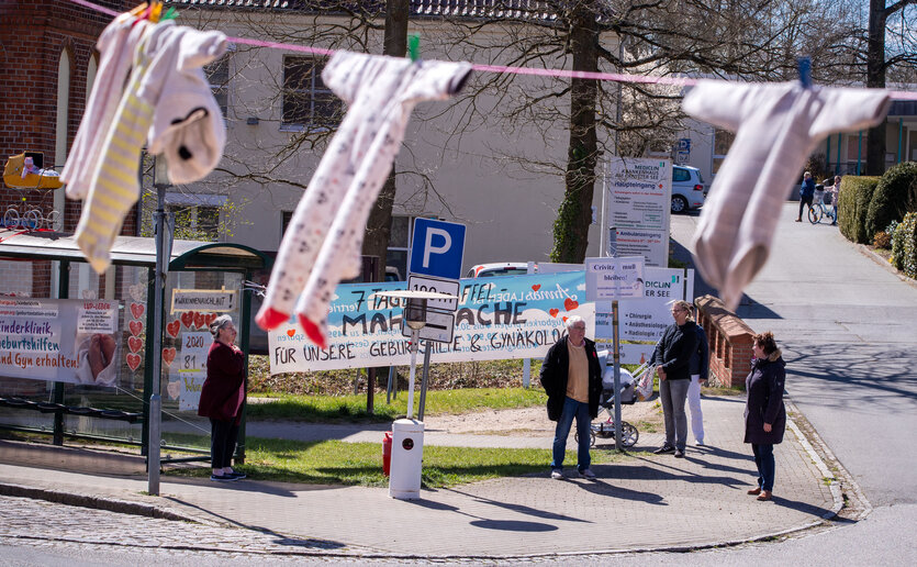 Im Vordergrund sind Babybodys an einer Wäscheleine aufgehängt, im Hintergrund stehen Menschen, die an einer Mahnwache teilnehmen