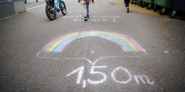 Auf einer Straße ist mit Straßenmalkreide ein Regenbogen gemalt worden. Darunter steht 1,50 Meter. Über dem Regenbogen sind Kinderbeine und ein Roller zu sehen.