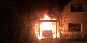 Das Auto von Ferat Kocak von hinten. Es steht in Flammen unter einem Carport neben einem Haus.