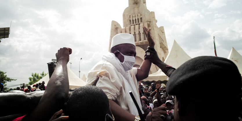 Imam Mahmoud Dicko in Mitten einer Menschenmenge. Er ragt über die Köpfe der andren empor und hebt seinen arm. Jemand berührt seine Hand. Dicko trägt eine Maske.