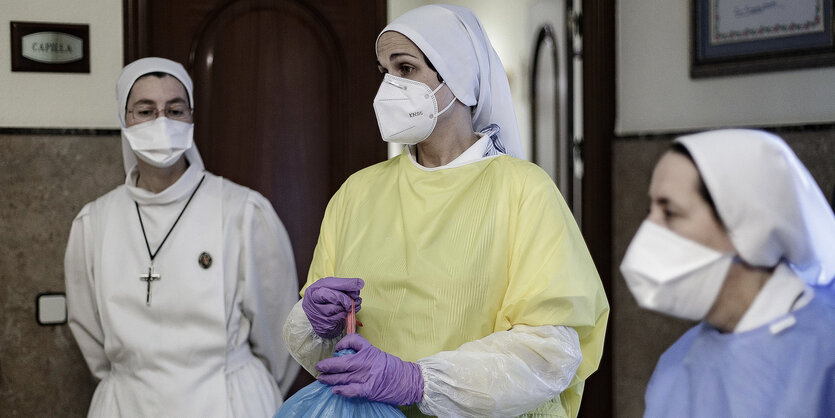 drei Nonnen mit Mundschutz und weißen Hauben. Die Nonne in der Mitte schnürt mit ihren Händen, über die sie lila Plastikhandschuhe gestreift hat, einen Müllsack zu