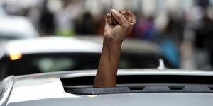 Der Arm einer schwarzen Person ragt aus dem Dachfenster eines Autos. Die Faust ist im protest geballt.