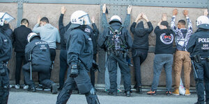 Eine Reihe junger Menschen mit erhobenen Händen vor einer Wand mit Eisenträgern, davor gepanzerte Polizisten