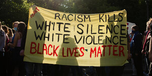 Ein Plakat mit der Aufschrift ·Racism kills, white silence, white violance, black lives matter· (Rassismus tötet, weißes Schweigen, weiße Gewalt, Schwarze Leben zählen) wird von Demonstrant*innen hochgehalten.