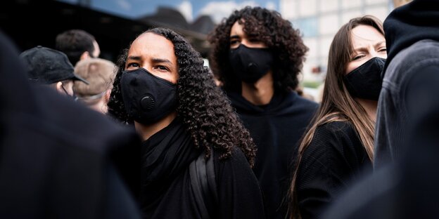 Schwarz gekleidete Personen mit Mundschutz stehen in einer Menschenmenge und protestieren