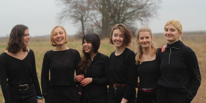 Sechs Frauen auf freiem Feld, schwarze Anziehsachen, ein Baum im Hintergrund