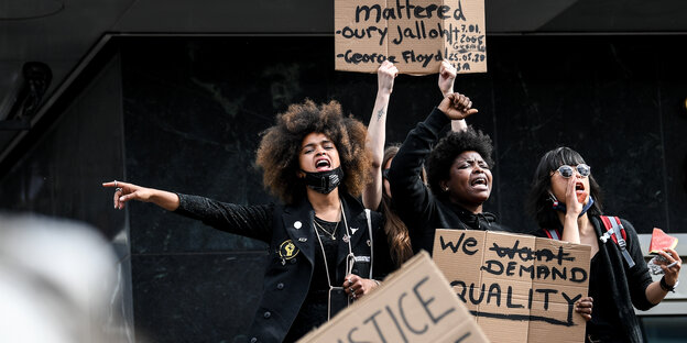 Drei schwarze Fraue halten Schilder hoch und rufen Sprechchöre auf einer Demo. Auf einem Schild steht: We demand Equality