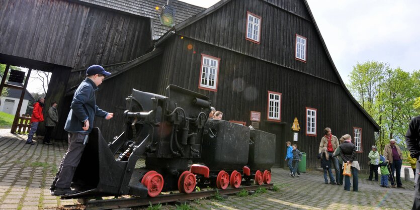 Eine Holzgebäude mit einer Grubenbahn im Vordergrund, neben der ein Junge mit Mütze steht, rechts im Hintergrund eine Familie