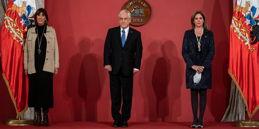 Zwei Frauen und ein Mann stehen auf einer Bühne mit Flaggen und rotem Hintergrund.