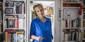 Eine blonde Frau mit Pferdeschwanz, die eine blaue Bluse trägt, steht in einem Türrrahmen, links und rechts der Tür sind Bücherregale