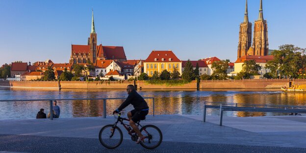 Ein Radfahrer fährt an einem Flussufer entlag, am anderen Ufer sind alte Gebäude