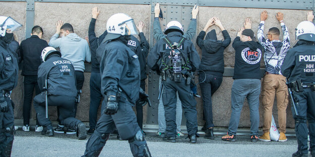 an einer Wand stehen Jugendliche, die die Hände hoch an die Wand halten, im Vordergrund läuft ein Polizist durch das Bild, er trägt einen Helm