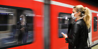 Eine Frau mit Mundschutz steht auf einem Bahnsteig neben einem einfahrenden Zug.