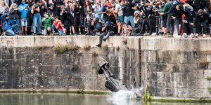 Eine Statue stürzt kopfüber ins Wasser, am Ufer stehen viele Menschen und beobachten das, einige fotografieren