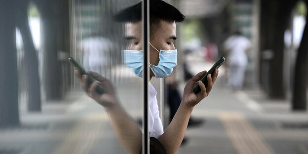 Mann mit Mundschutz schaut auf sein Smartphone, er wird von einer Glasscheibe gespiegelt