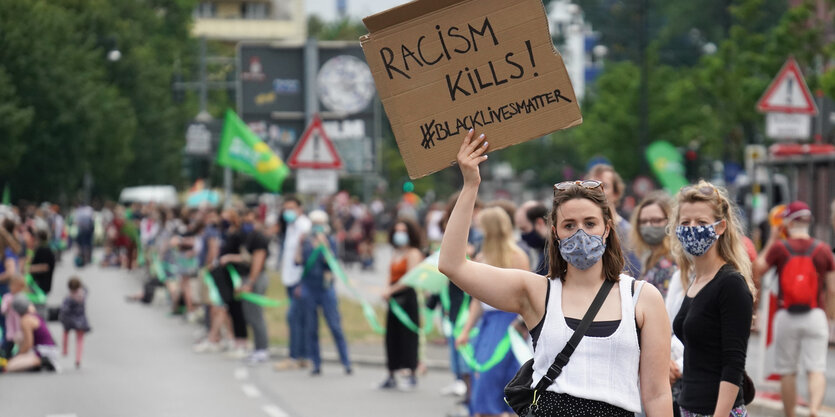 Ein Frau hält ein Plakat mit der Aufschrift Black-Lives-Matter hoch, dahinter sieht man menschen, die ein Band halten