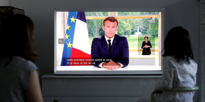 Zwei Personen schauen auf einen Bildschirm, darauf ist Emmanuel Macron zu sehen