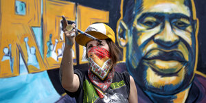 Künstlerin Greta McClain mit Pinsel in der Hand vor einem Wandgemälde, das einen stilisierten George Floyd zeigt.