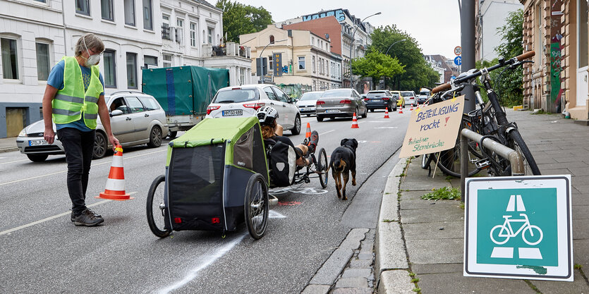 Ein Mann mit Warnweste stellt Hütchen auf einer Fahrbahn auf, neben ihm ein Fahrradanhänger