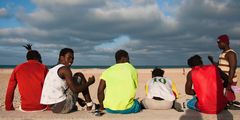 Menschen sitzen am Strand, sie tragen bunte T-Shirts, man sieht sie von hinten