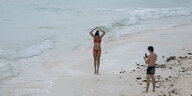 Frau in rotem Bikini springt in die Luft am Meer, ein Mann schaut in sein Smartphone
