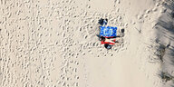 Ein nacker Mensch liegt im Sand auf einem Handtuch, viele Fussspuren im Sand