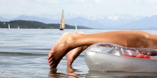 Füsse hängen über eine Luftmatratze in den See, ein Segelboot und Berge im Hitnergrund