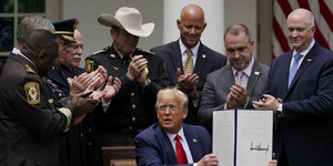Donald Trump hält eine Mappe hoch, auf der seine Unterschrift zu sehen ist. Um ihn herum stehen Männer, viele davon uniformiert.