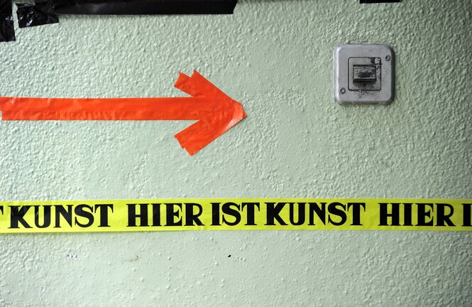 Ein ornagener Pfleil und ein gelbes Klebeband mit dem schwarzen Schriftzug "Hier ist Kunst" weisen auf eine kleine Galerie in der Neuköllner Wissmannstraße hin