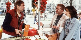 Harald Juhnke steht an einem Marktstand und hobelt Gemüse mit einem Gemüsehobel, vor ihm stehtn zwei jüngere Leute