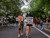 Zwei Frauen mit Mundschutz auf einer Straße bei einer Demonstration, eine hält das Schild "Defund the police" hoch. Zwei Frauen mit Mundschutz auf einer Straße bei einer Demonstration, eine hält das Schild "Defund the police" hoch.