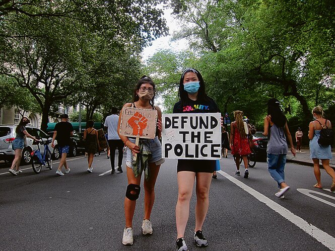 Zwei Frauen mit Mundschutz auf einer Straße bei einer Demonstration, eine hält das Schild "Defund the police" hoch.