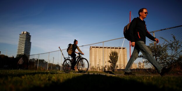 Fahrradfahrerin und Spaziergänger auf einem Weg, dahinter ein Zaun und ein Industriegebäude